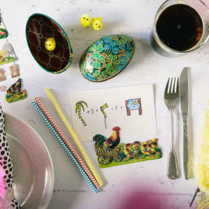 Ett bord fullt med påskrelaterade prylar: ett kort med en tupp, två påskägg, ett glas med mjöd och några påskkvistar.