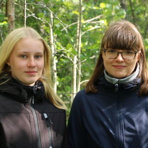 En blond tjej och en med mörkt hår står i en skog.