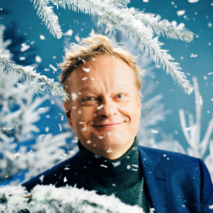 Pianisti Iiro Rantala katsoo puolilähikuvassa hymyillen kameraan, ympärillään luminen kuusi ja sen "hiutaleita".