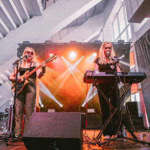 Vaaleahiuksinen nainen aurinkolasit päässään soittaa kitaraa lavalla, toinen vaaleahiuksinen nainen soittaa syntetisaattoria. Taustalla on savua ja valkeaa valoa.