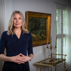 En blond kvinna med mörkblåklänning tittar allvarligt in i kameran. Hon har händerna knäppta. Bakom henne syns en tavla med guldram.