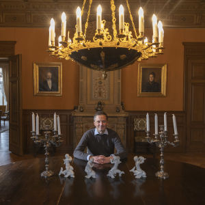 Henrik Creutz har händerna knäpta och sitter vid ett stort matbord. Takkronan hänger ovanför honom.