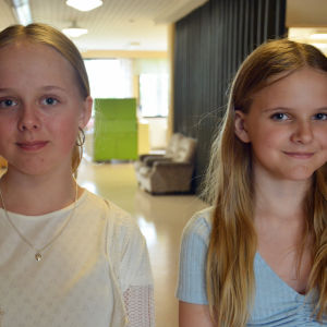 Två flickor i 11-årsåldern står i en skolkorridor och tittar in i kameran.