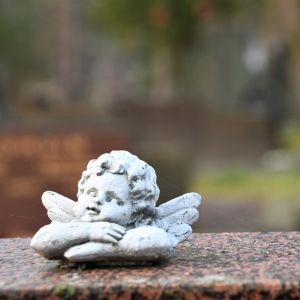 En vit kerubaktig ängel sitter med armarna i kors på en gravsten av marmor. 