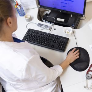 En läkare sitter vid datorn och skriver och mittemot sitter en patient.