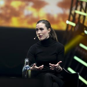 Sanna Marin svardklädd med en mikrofon på en scen, hon sitter och håller händerna lyfta med handflatorna uppåt.