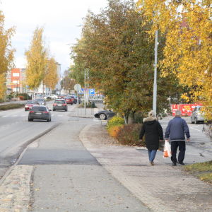 Ett äldre par går på gatan. På vägen till vänster om paret kör flera bilar.
