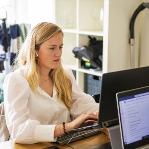 En kvinna sitter vid ett bord och jobbar på en dator. I bakgrunden hänger tvätt på en bykställning.