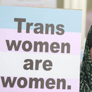 En sminkad demonstrant i huvudduk med med en skylt med texten "Trans women are women", alltså "Transkvinnor är kvinnor".