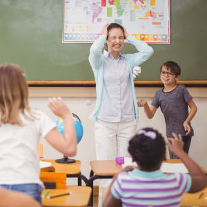 Kuvassa on päätään pitelevä opettaja luokassa. Oppilaat ovat selin kuvaan ja heittelevät paperipalloja opettajaa kohti. 