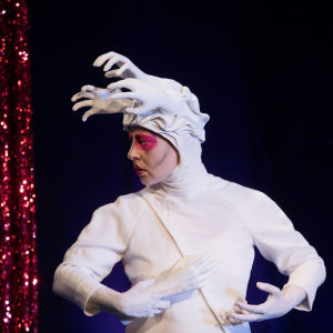 Drag-artisti Theodora Rex pukeutuneena valkoiseen asuun, jonka hupussa valkoisia tekokäsiä.