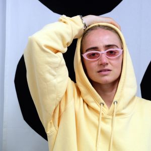 Den svenska hiphop-artisten Silvana Imam sitter med ena handen bakom huvudet, hon har på sig glasögon och en gul munkjacka.