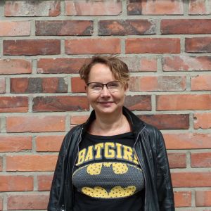 Tutkija Tuula Juvonen kuvattuna tiiliseinän edessä. Hänen päällä on nahkatakki ja t-paita, jossa teksti "batgirl".