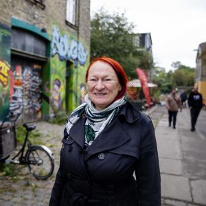 Kirsten Larsen tittar in i kameran och ler. Hon står på en gata i Christiania.