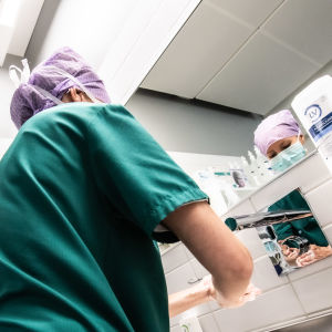 En kvinna med håret bakbundet och skyddat tvättar händerna innan hon går in i en operationssal. Hon har på sig gröna kirurgkläder.