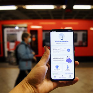 En hand håller upp en telefon med appen Coronablinkern, på en metroperrong.