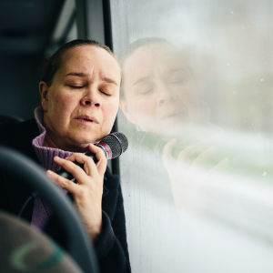 Katja Küttner istuu bussissa ja nojaa uupuneena, silmät kiinni ikkunaan. Kädessään hänellä on mikrofoni.