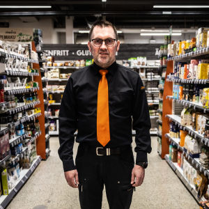 Man i mörk skjorta och orange slips står mellan butikshyllor