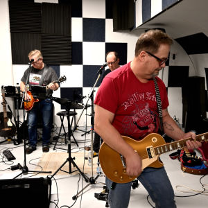 En man i röd t-skjorta spelar elgitarr, i bakgrunden en sångarska och två gitarrister