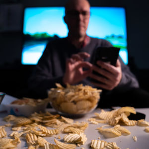 Epätarkka mieshahmo räplää kännykkää television äärellä, pöytä kuorrutettuna sipseillä ja muilla roskaruoilla.