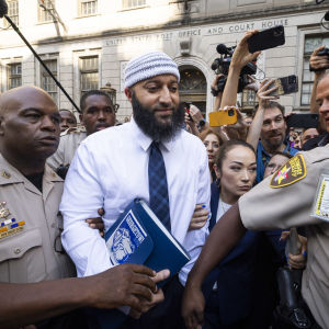 En skäggig man i vit-blå-randig mössa, blå slips och vit skjorta med en bok i handen går med poliser genom en folkmassa.