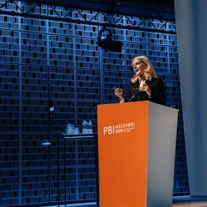 A woman speaking behind an orange PBI Helsinki 2019 podium in front of a blue wall.