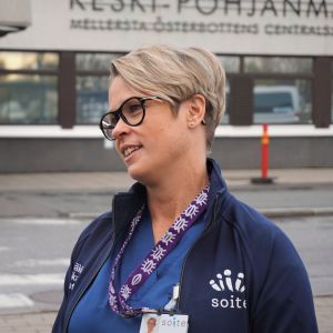 Ljushårig kvinna med glasögon står framför ingången till ett sjukhus.