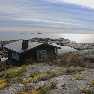 Pieni ruskea mökki sinisillä ikkunanpielillä kalliosella saarella ulkosaaristossa. Kallioiden takaa näkyy meri.
