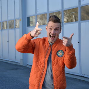 Programledaren Jontti Granbacka står på en parkering iklädd en orange jacka med logon för nya barnprogrammet Superhjälparna.