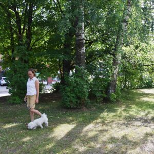 Kvinna i topp och shorts går i en grön park tillsammans med sin hund. Bakom buskarna och träden skymtar höghus.