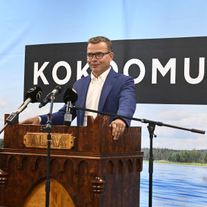 Petteri Orpo står och håller tal vid partimötet i Seinäjoki, bakom sig har han ett sjölandskap. 