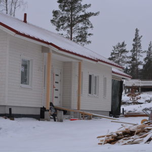 ett halvfärdigt hus, husbygge på gång. Vinter och snö. En hög med bräden. I bakgrunden ett färdigt, nybyggt hus.