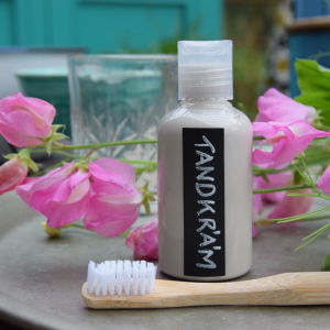 En plastflaska med hemgjord tandkräm och en tandborste av bambu. I bakgrunden en luktärt.