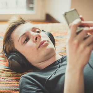 En kvinna med kort hår ligger på en säng och lyssnar på musik. Hon har hörlurar på sig och tittar på sin smarttelefon. 