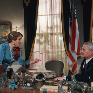 Märta står vid ett stort skrivbord och pekar bestämt mot presidenten som sitter i stolen.