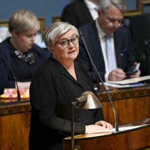 Anu Vehviläinen i mörka klädet står och talar i riksdagen, i bakgrunden syns bland annat Pekka Haavisto och Annika Saarikko.