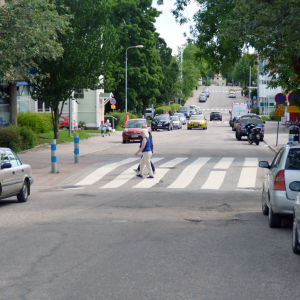 Några personer går över ett övergångsställe vid en väg där många bilar har parkerat.