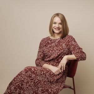Kuvassa on kirjailija Meri Valkama. Hän istuu tuolilla ja hymyilee kameralle. 