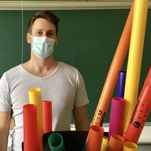 Man med munskydd  står framför svarta tavlan i klassrum, framför sig har han färgglada plaströr.