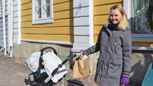 Annukka Kokki. En ung kvinna står och håller i handtaget till en barnvagn. Bakom henne synns en gul husvägg.