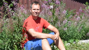 Jan Glantz njuter av semester i Västnyland. Han jobbar och bor numera i Helsingfors. en man i röd t-skjorta och blå shorts sitter på en låg betongmur. Han omgärdas av duntrav, det vill säga rödlila blommor.