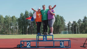 Kolme naista tuulettavat palkintokorokkeella urheilukentällä, kaulassaan mitalit.