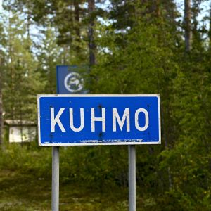 En trafikskylt med texten "Kuhmo" och skog i bakgrunden.