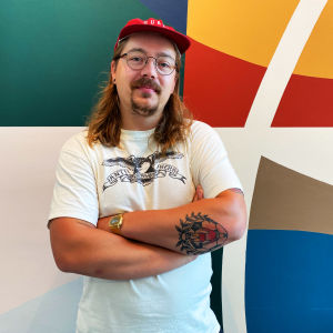 En glasögonprydd Kent Ketomäki iklädd t-skjorta och keps står med armarna i kors framför en färggrann vägg.