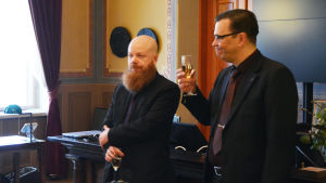 Borgå stadsstyrelses orförande Jaakko Jalonen skålar för Kännö. Två män står och håller i glas med skumvin framför andra människor.
