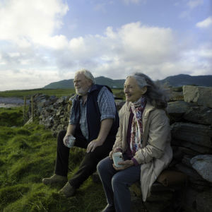 En leende Howard (James Cosmo) och Annie (Bríd Brennan) sitter vid en stenmur och ser ut över havet, bakom dem syns gröna vidder och berg.