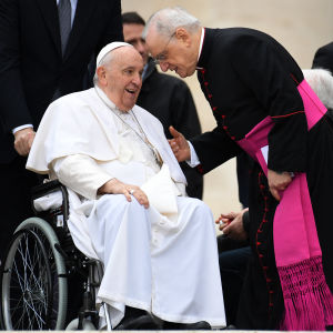 Tummaan pukeutunut mies puhuu pyörätuolissa istuvalle Paavi Franciscukselle nojautuneena eteenpäin.