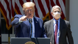 USA:s president Donald Trump och smittskyddsexpert ANthony Fauci är glada över att ett nytt vaccin kan tas i bruk. Donald Trump står vid ett talarpodium och håller presskonferens. I bakgrunden syns smittskyddsexperten Anthony Fauci som bär ansiktsskydd. Trump är utan.
