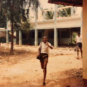 Hymyilevä nuori srilankalainen poika kävelee kohti kameraa koulun raunioiden edessä.