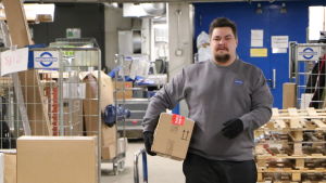 Mathias Rosenlund går i en logistikcentral iklädd arbetskläder med ett paket under armen. 